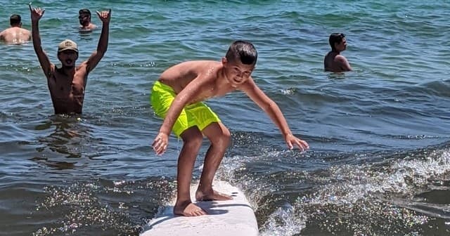 Child balancing on surfboard with swimmers cheering in shallow sea