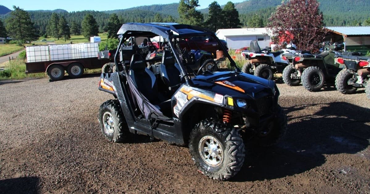 Two-seat UTV parked on dirt trail near trailers and pine trees
