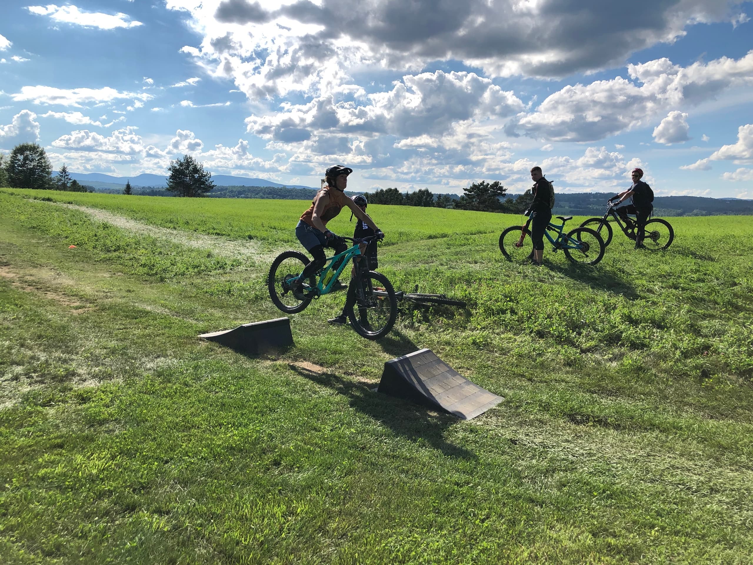 Mountain bikers riding grassy hill with jump under blue sky