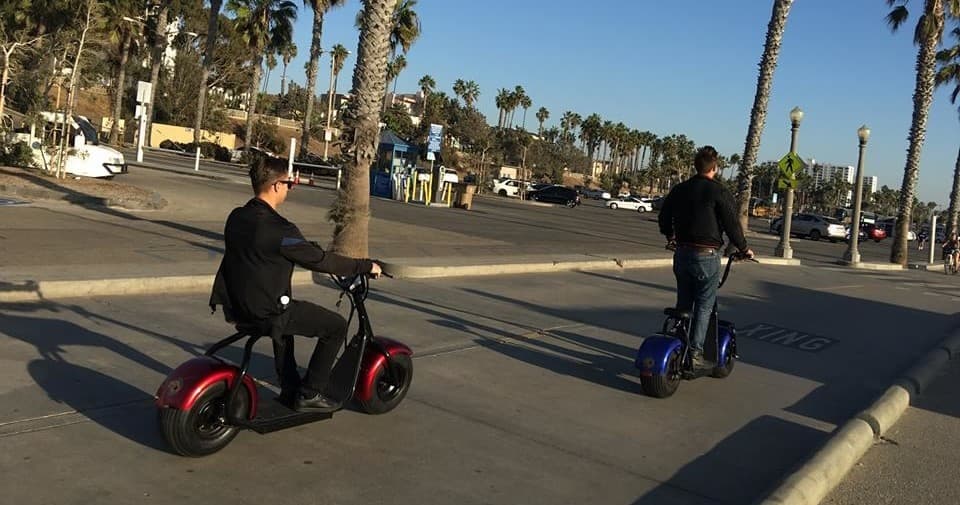 Two riders cruising coastal boardwalk by palm trees on e-scooters