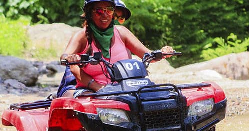 Female rider on red ATV splashing river rocks, outdoor trail