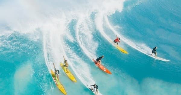 Aerial of multiple surfers racing down turquoise open ocean waves