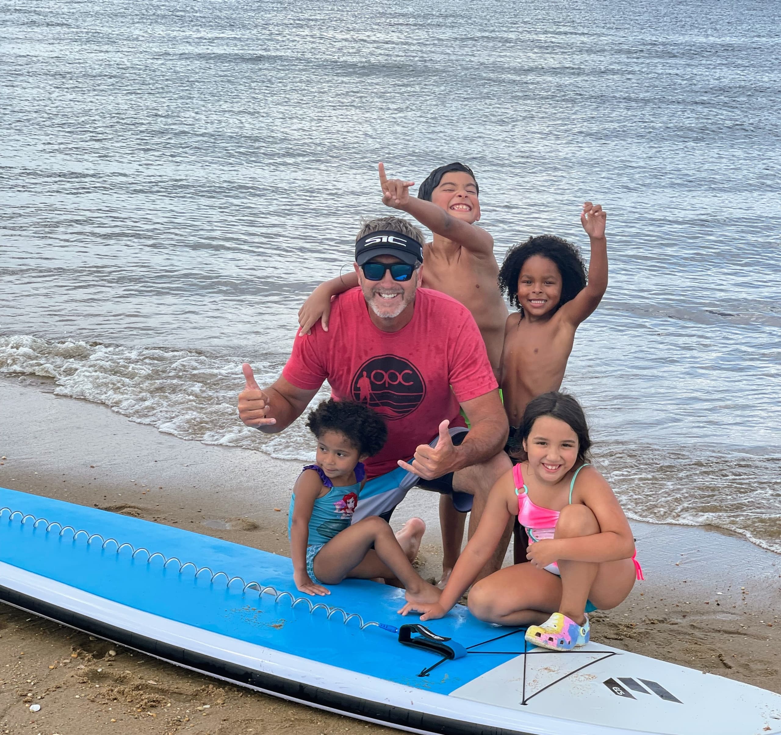 Family posing on beach with paddleboard, kids playing at shoreline