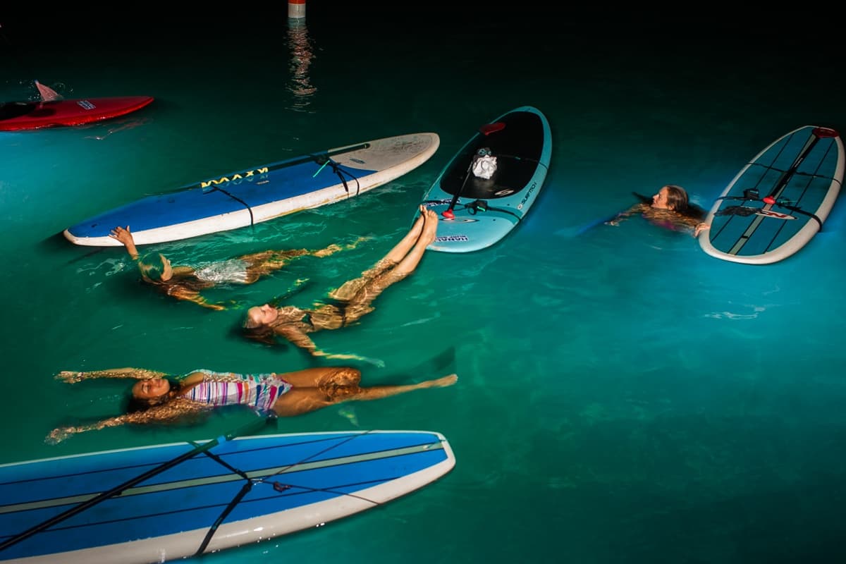 People floating on paddleboards in glowing turquoise night bay