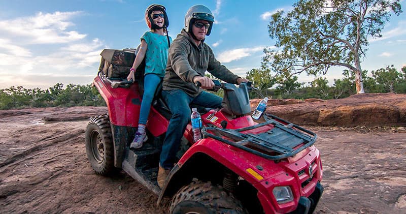 Couple on red quad bike crossing rocky outback with scenic sky