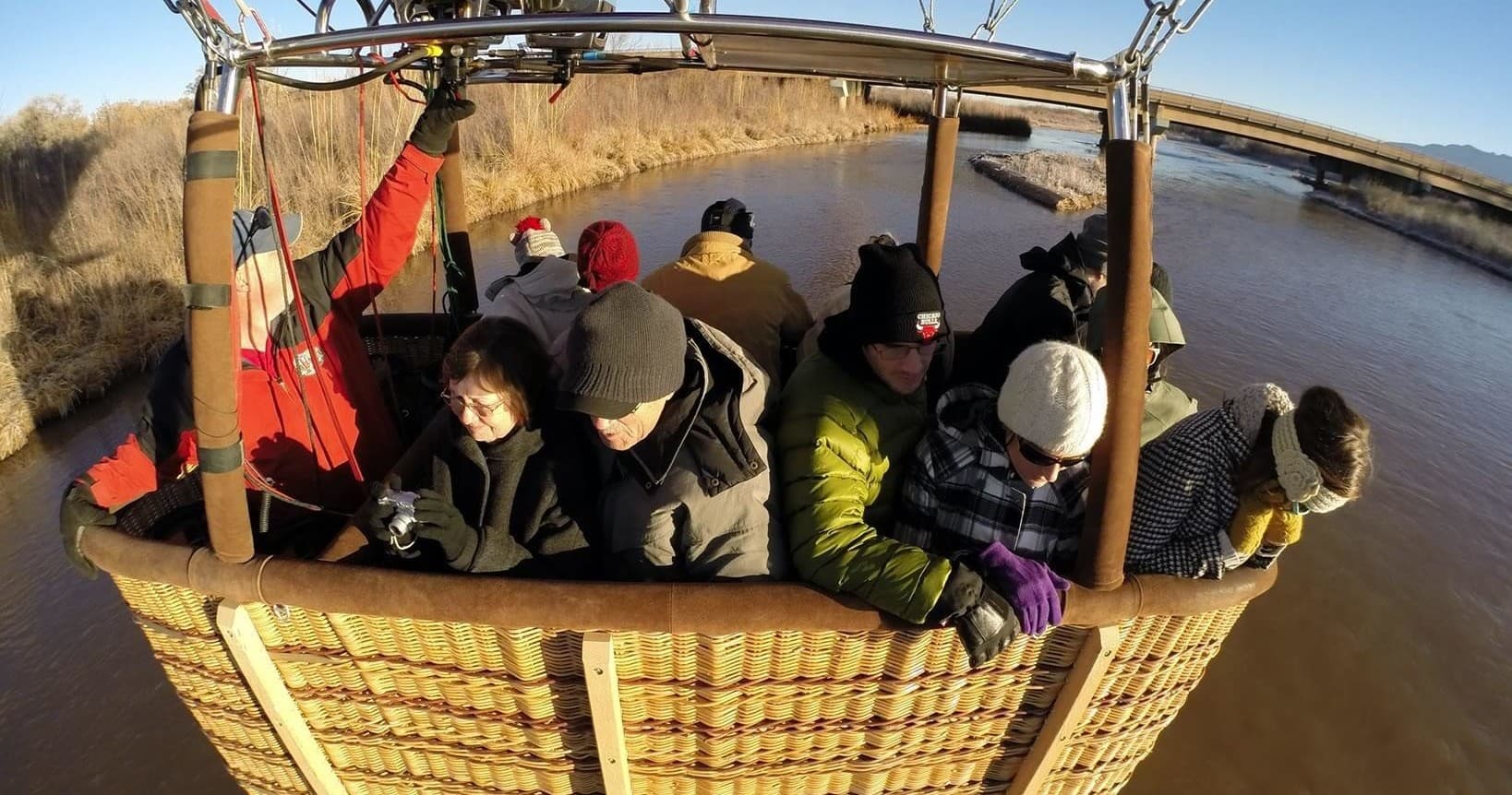 Passengers smiling in hot air balloon wicker basket above riverbanks