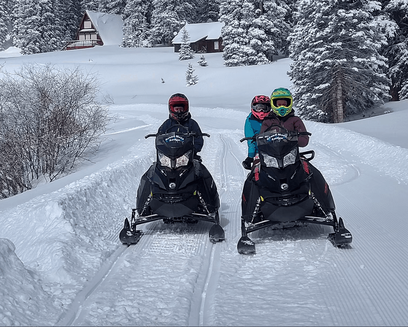 Two people snowmobiling on groomed snowy trail in alpine forest