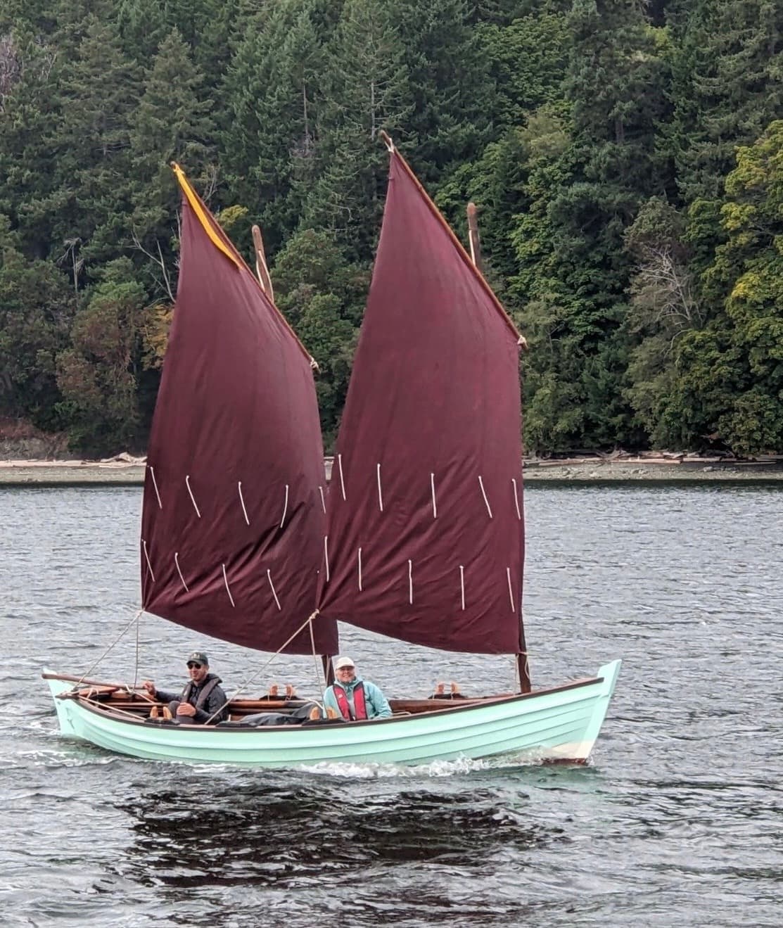 Traditional two-masted sailboat near forested shoreline on water