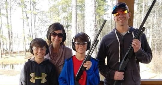 Family at outdoor shooting range holding rifles near woods