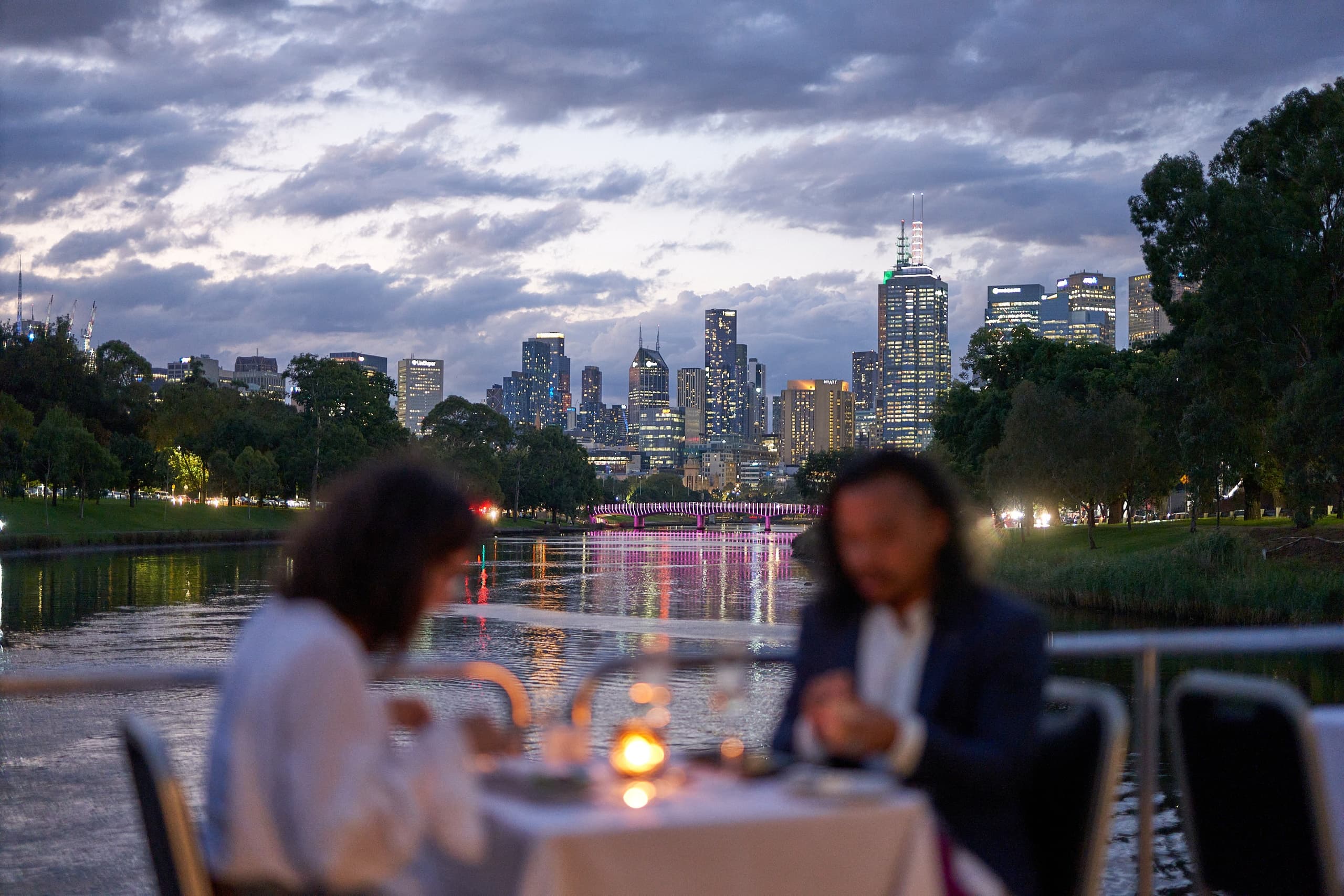 Couple dining by Yarra River at dusk with illuminated Melbourne skyline