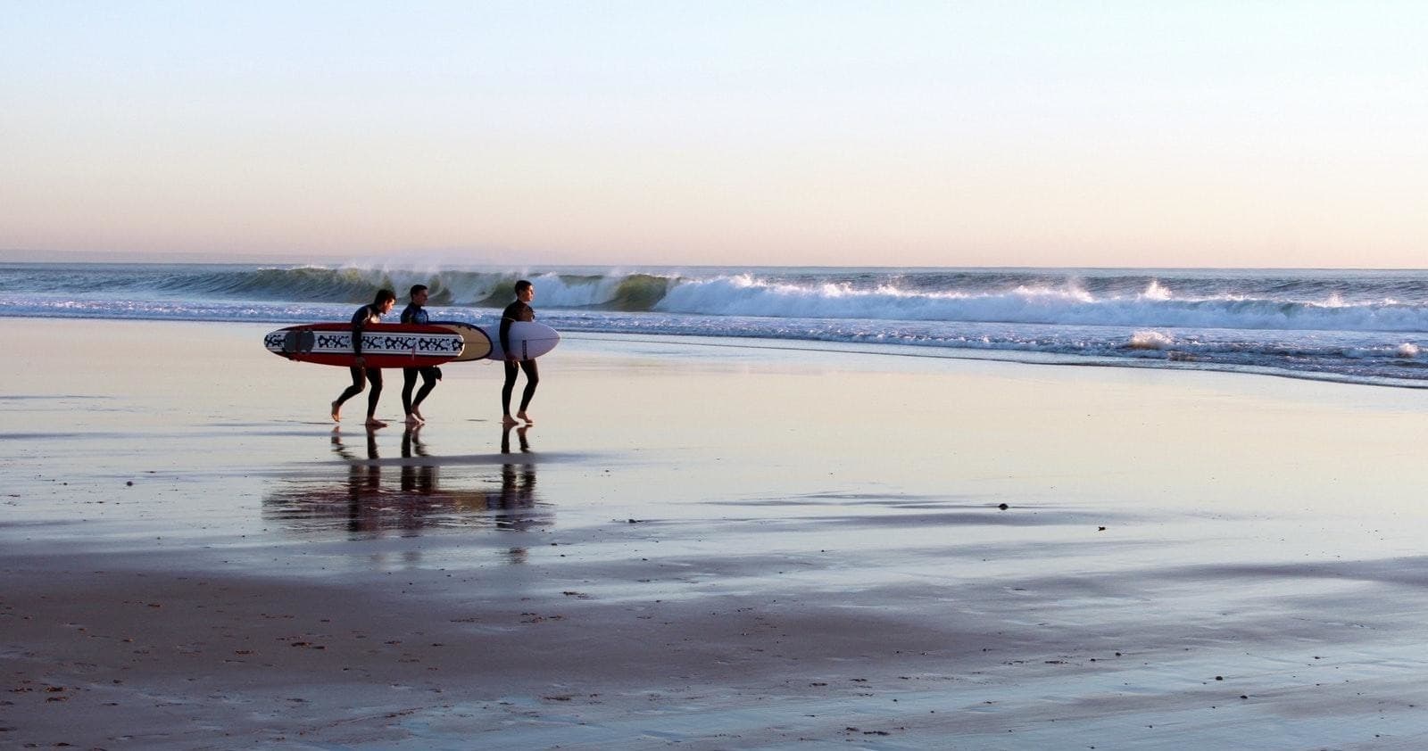 Three surfers carrying boards along reflective shoreline at sunset