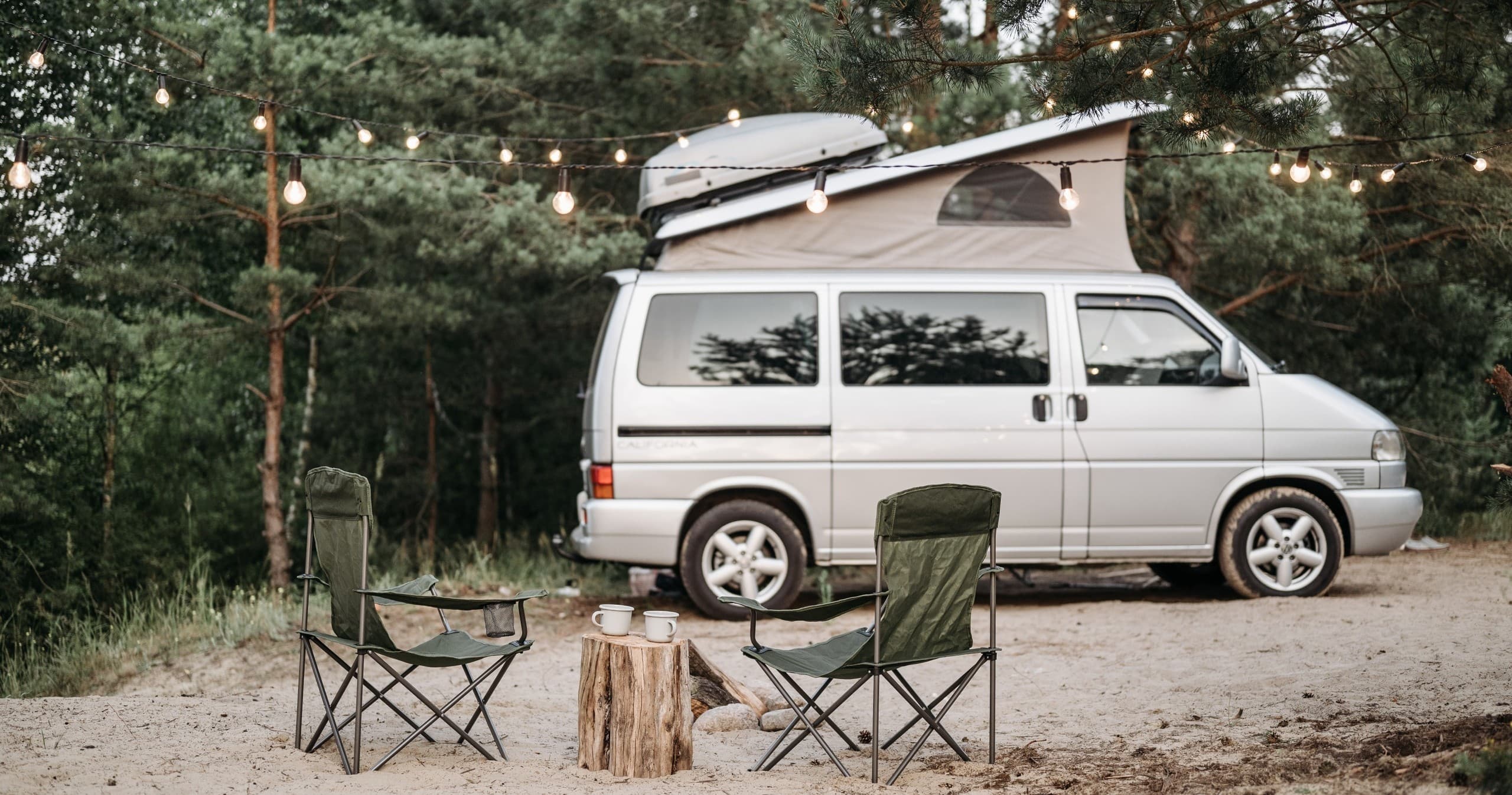Camper van campsite in forest clearing with chairs and lights