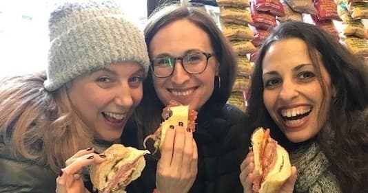 Three friends eating sandwiches on Boston food tour, smiling