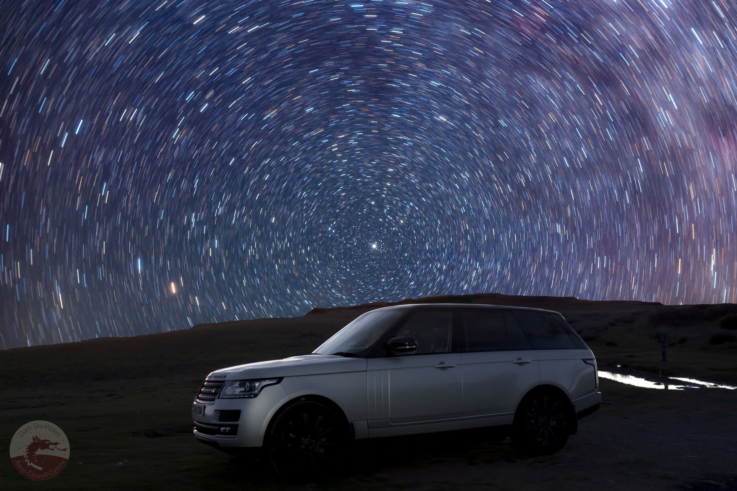 SUV parked under rotating star trails across rural night sky
