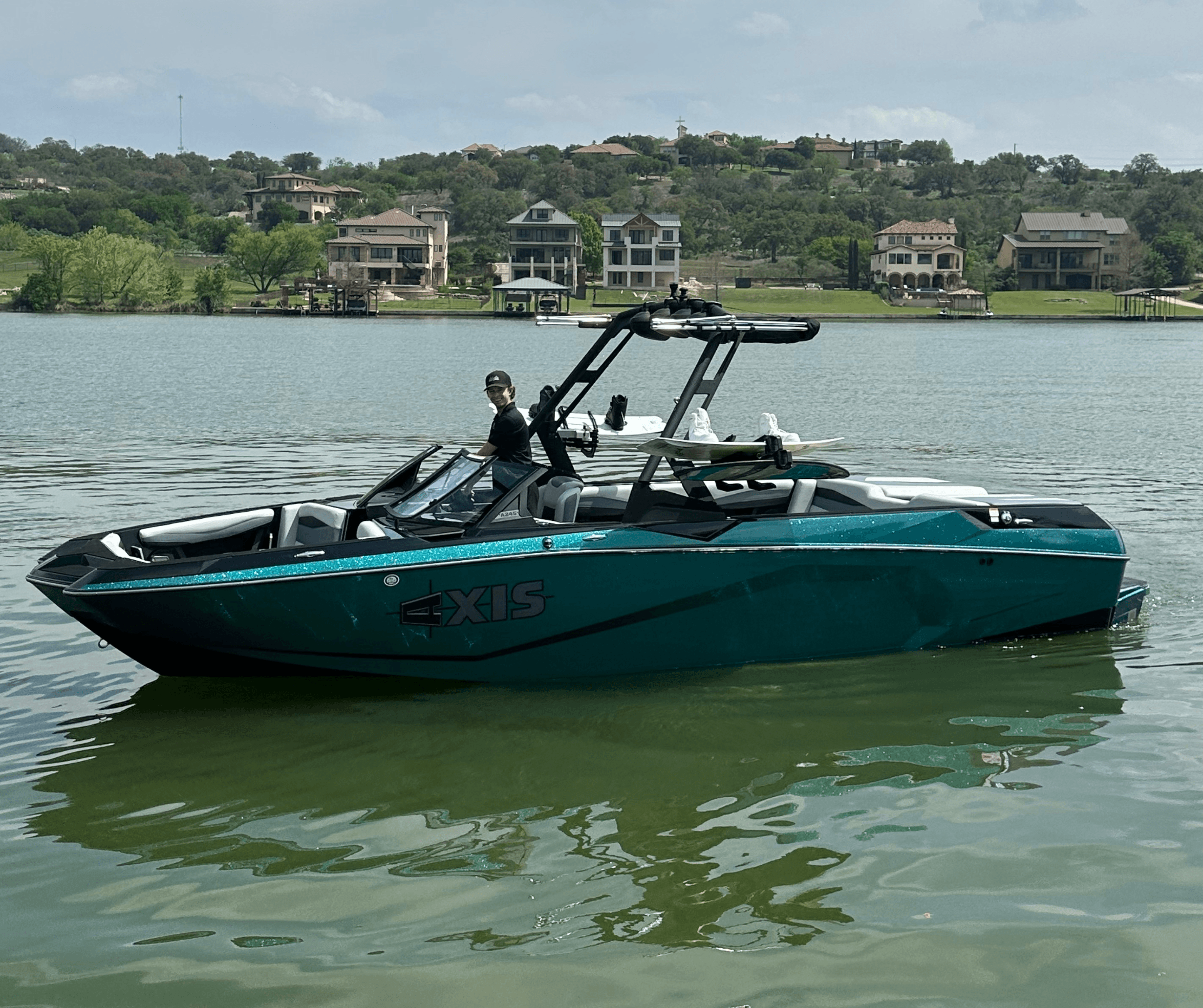 Teal wake/surf boat cruising on a calm lakeside near homes
