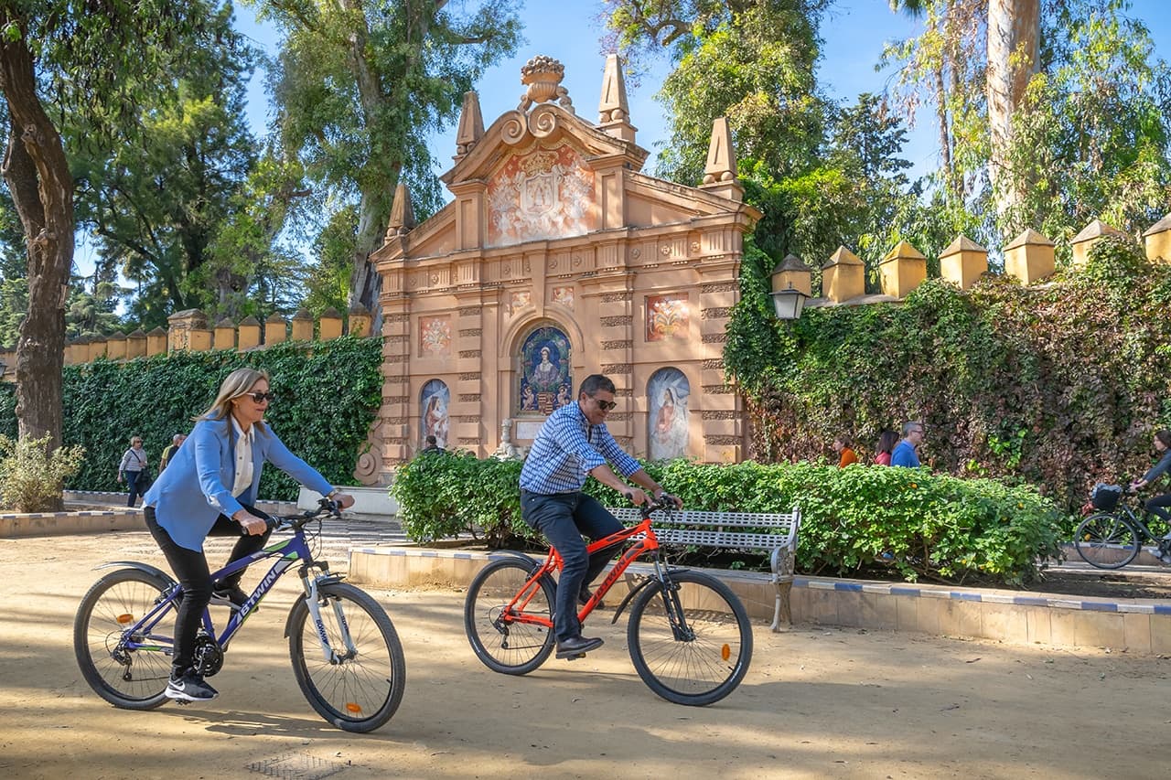 Two cyclists pedaling past ornate park gate under blue sky