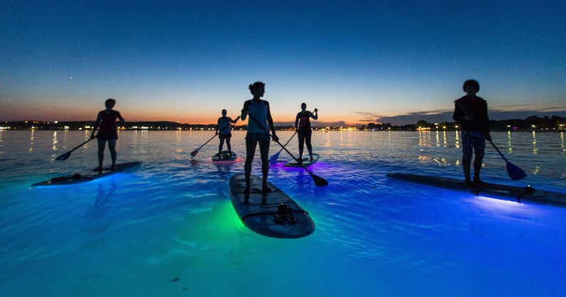 Glow paddleboarders on water at twilight with LED lights