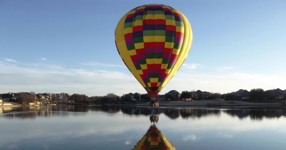 Hot air balloon reflected over calm lake shoreline at sunrise
