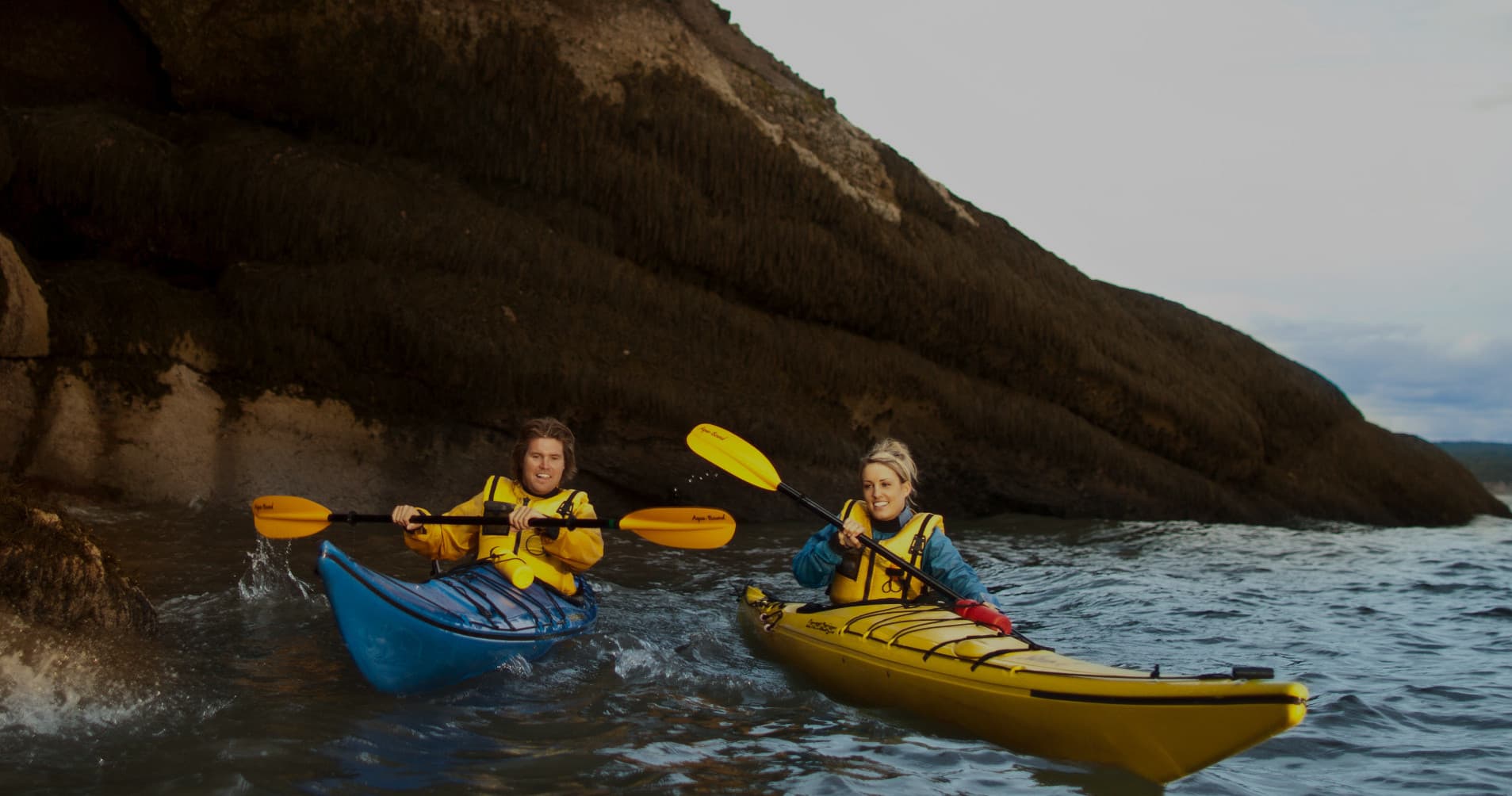 Two kayakers paddling along rocky coastal shoreline at dusk