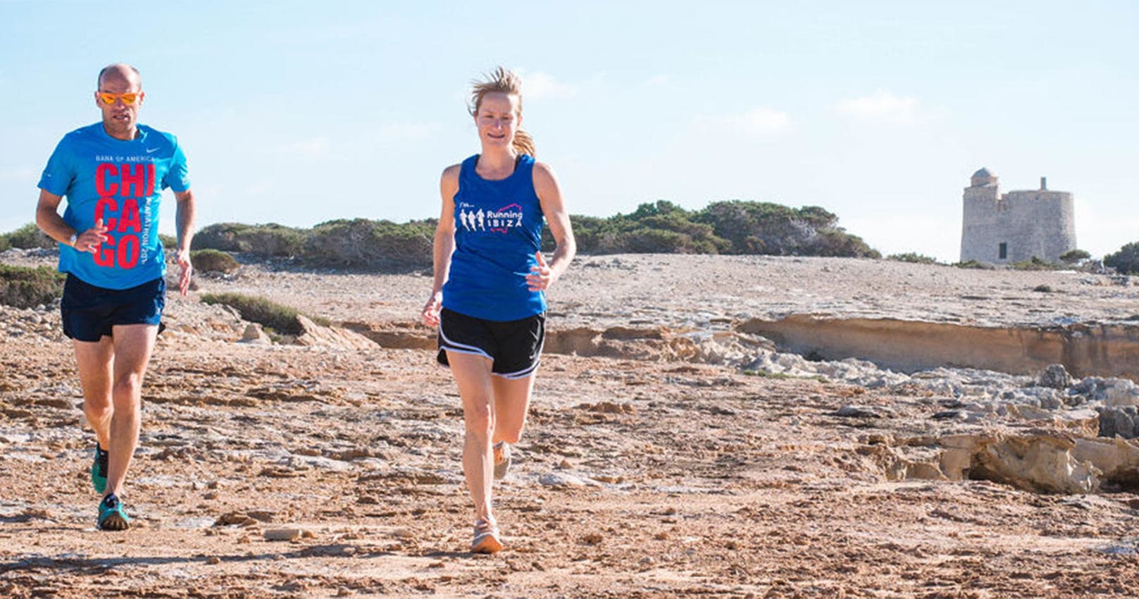 Runners jogging along rocky Ibiza coastline near watchtower