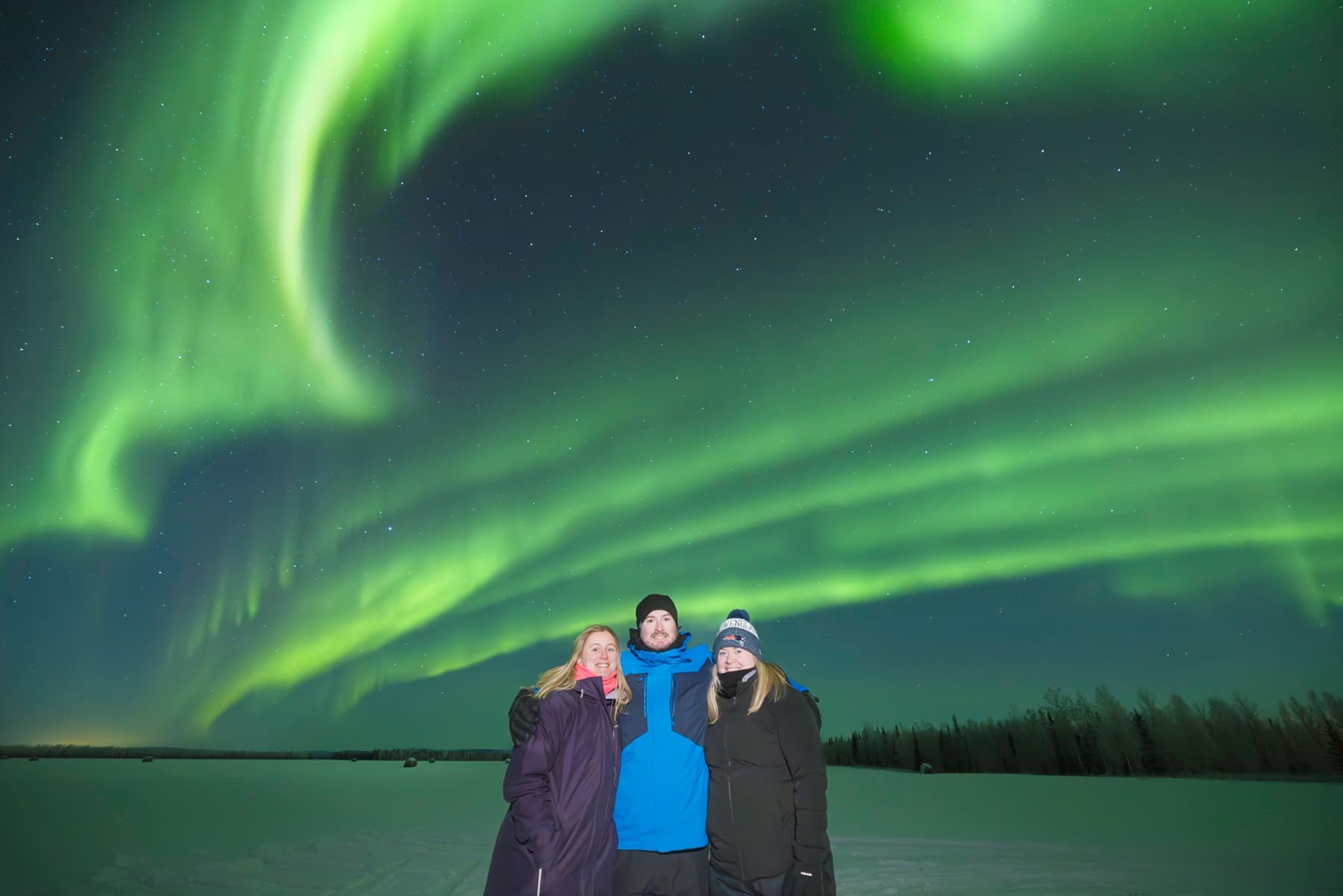 Three people viewing vivid northern lights over frozen lake
