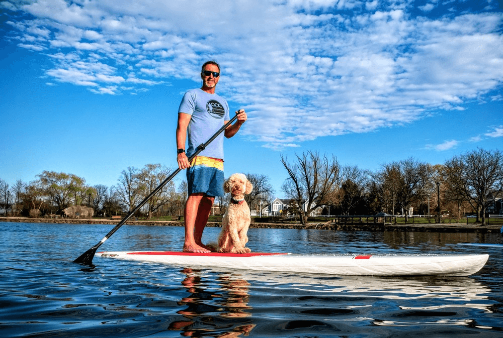 Stand-up paddleboarder with dog on calm lake under blue sky