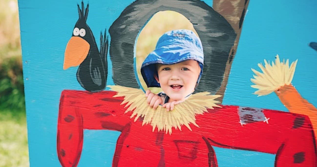 Child peeking through scarecrow cutout at outdoor farm fair