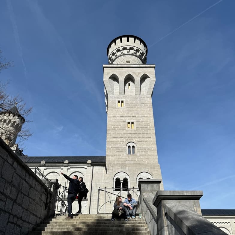 Visitors on stone steps beside a tall castle tower under blue sky