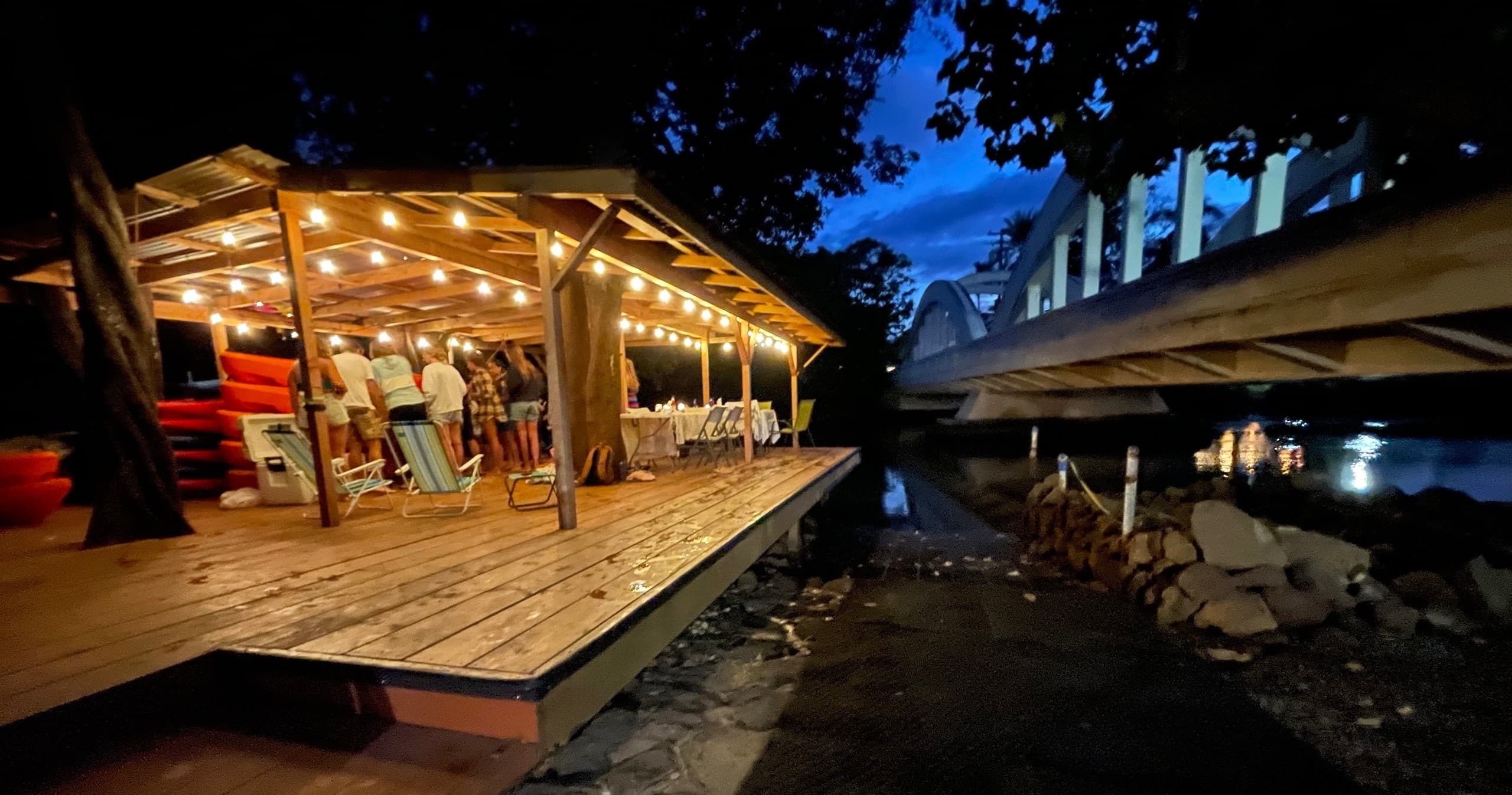 Riverside lit wooden deck dining under bridge at dusk