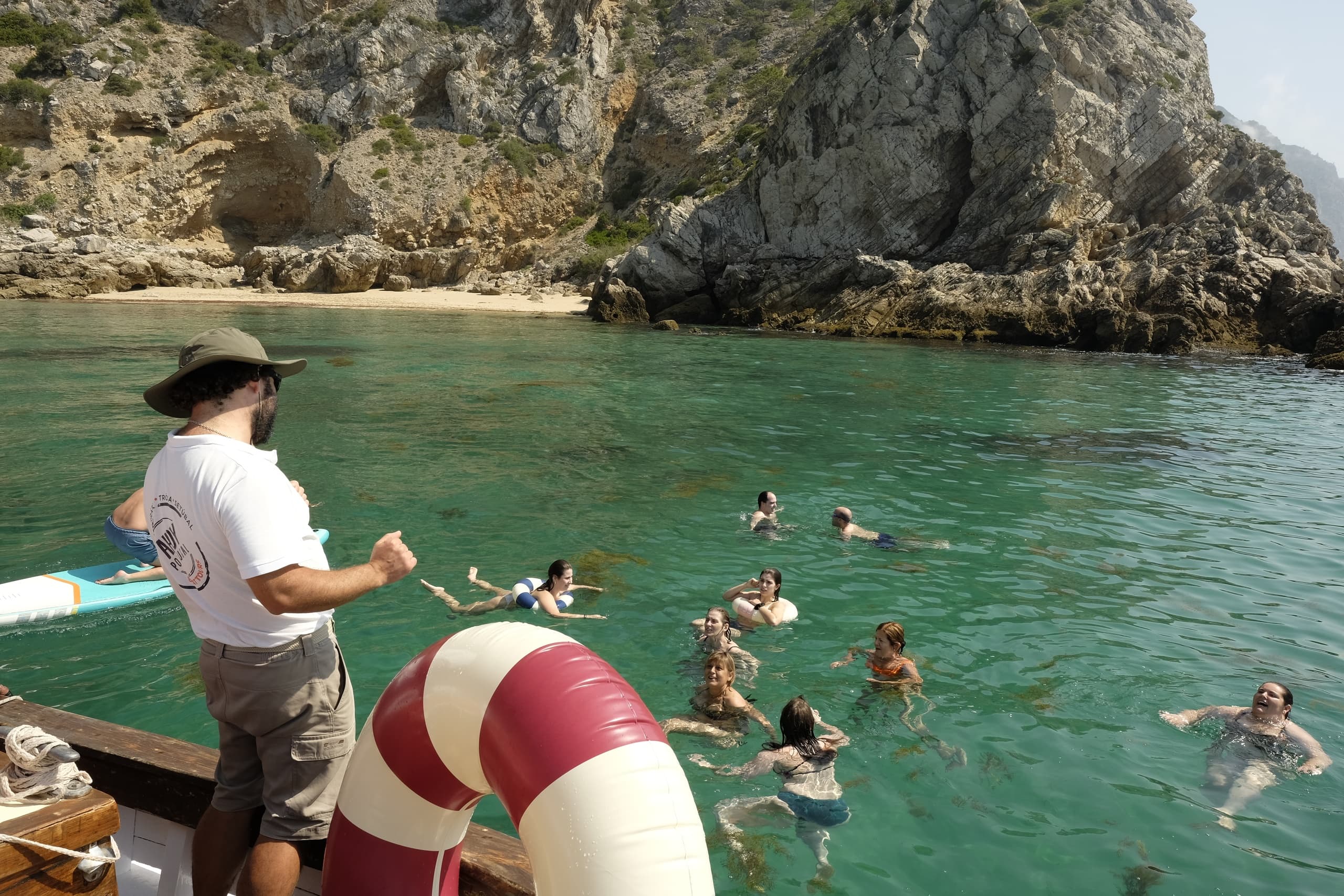 People swimming from sailboat in turquoise cove by rocky coast