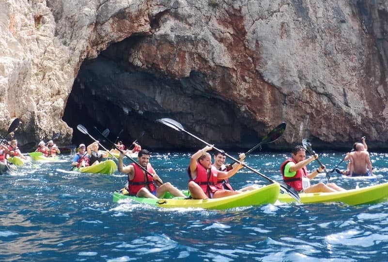 Group kayaking along rocky sea caves under bright Mediterranean sky