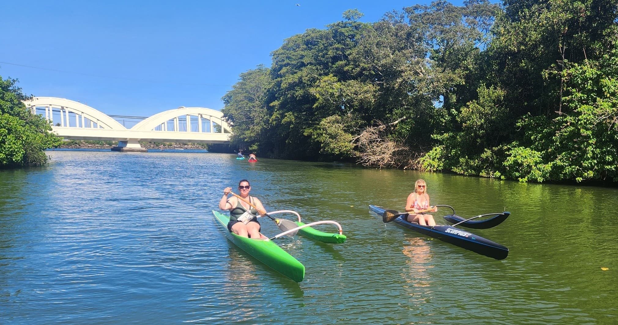 Two kayakers paddling a scenic river with bridge and green banks, Hawaii