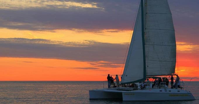 Catamaran sailing at sunset on open ocean, passengers silhouetted