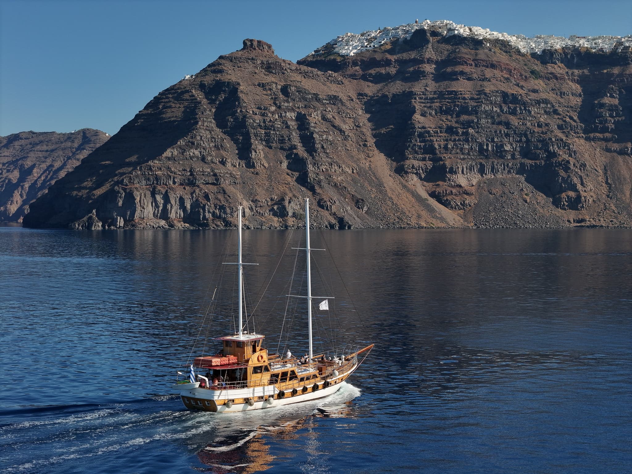 Traditional wooden yacht sailing near Santorini cliffs, daytime cruise