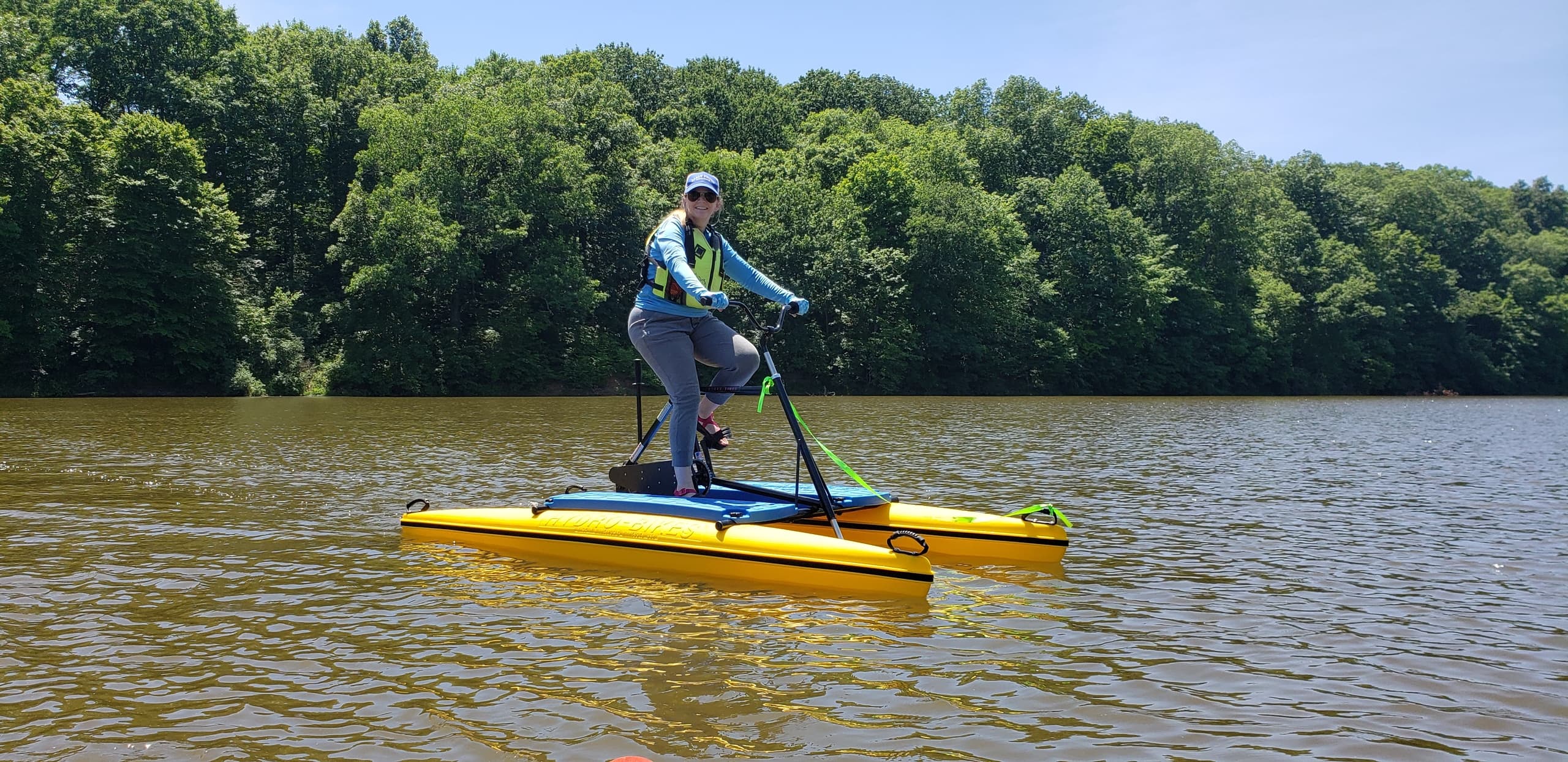 Person paddleboarding on twin-hull SUP across calm river, wooded bank
