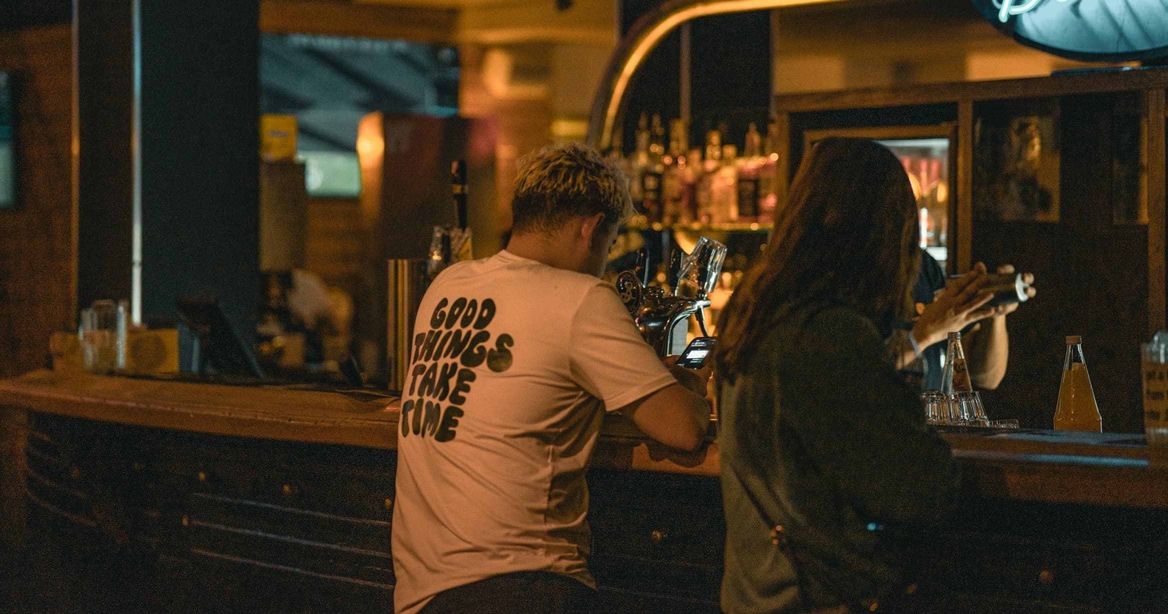Two people seated at pub bar counter ordering drinks in warm lighting