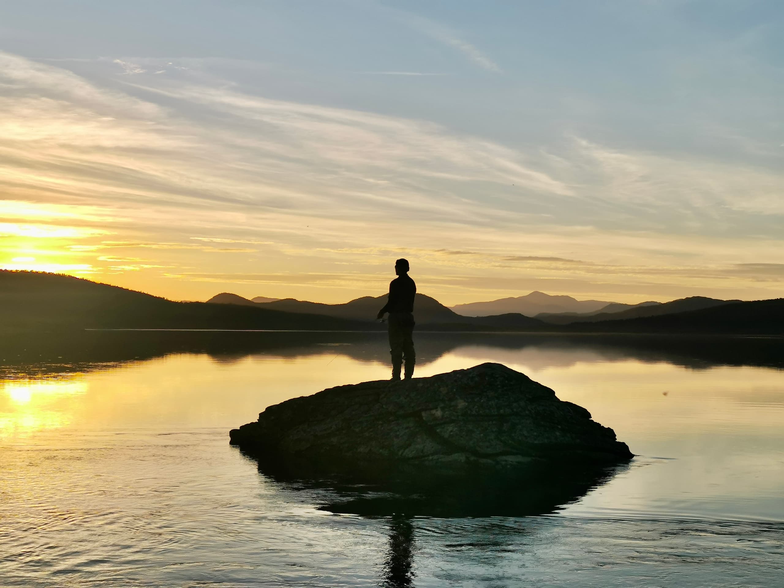 Hiker silhouetted on rock at sunset over calm Arctic lake
