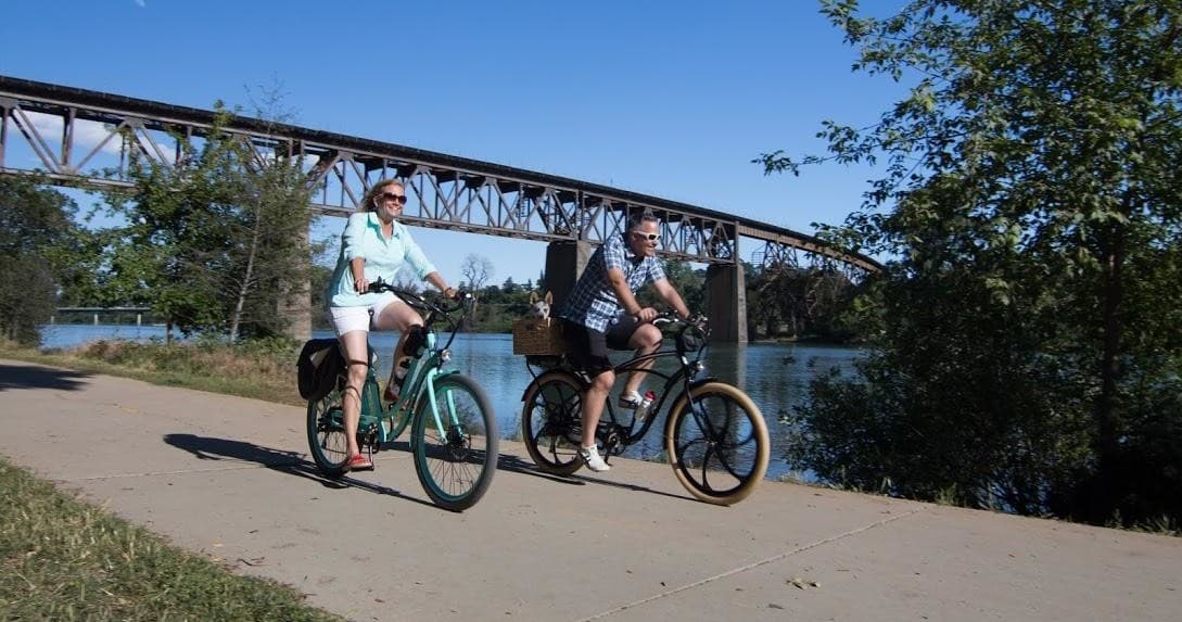 Two cyclists on electric bikes along scenic river path under bridge