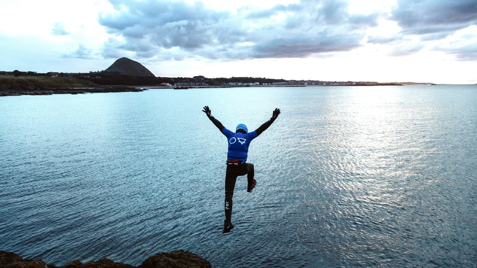 Wetsuit jumper leaping into calm ocean from rocky shoreline