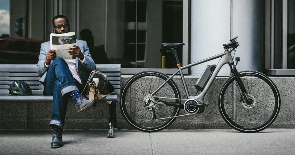 Man reading newspaper seated beside e-bike on urban sidewalk