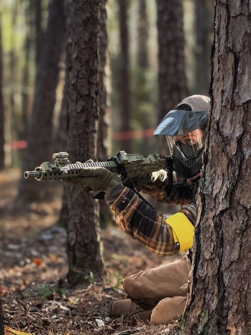 Player kneeling behind tree aiming airsoft rifle in pine woods