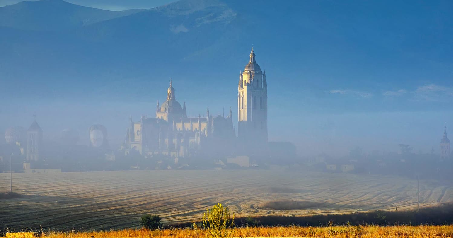 Misty Segovia cathedral rising from fields at sunrise, wide vista
