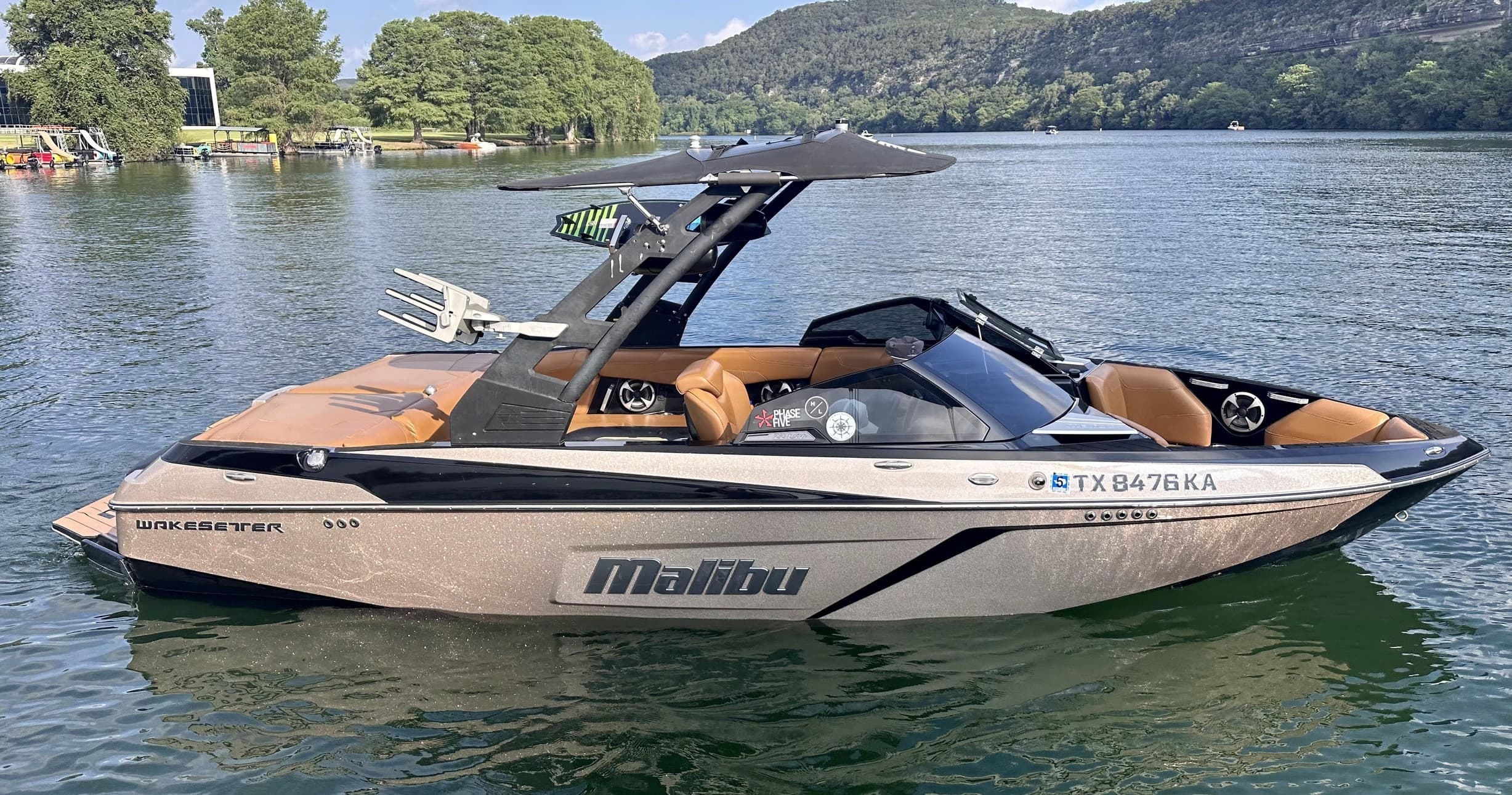 Tan Malibu wake boat floating on calm lake by green hills