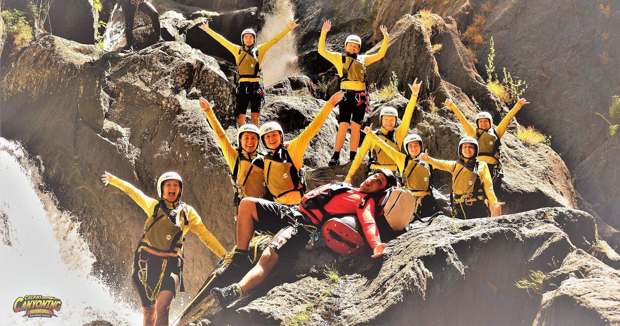 Group canyoning team celebrating on rocky waterfall ledge in rainforest