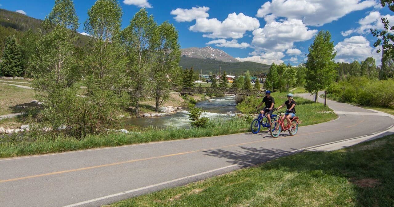 Cyclists riding paved riverside trail with mountains and blue sky