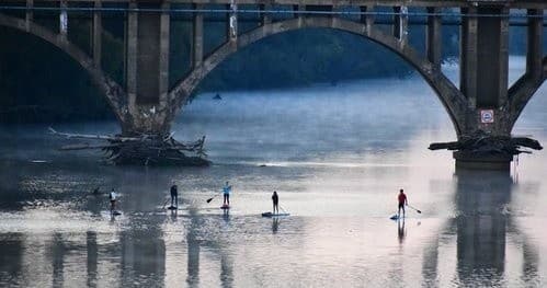 Standup paddleboarders gliding under misty stone bridge