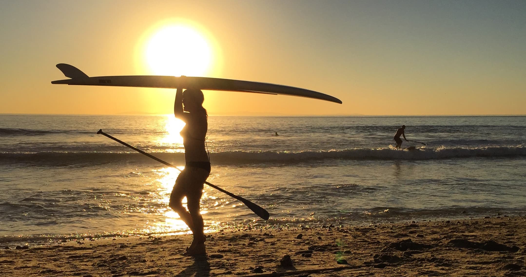 Silhouetted paddleboarder carrying board on sunset beach shore