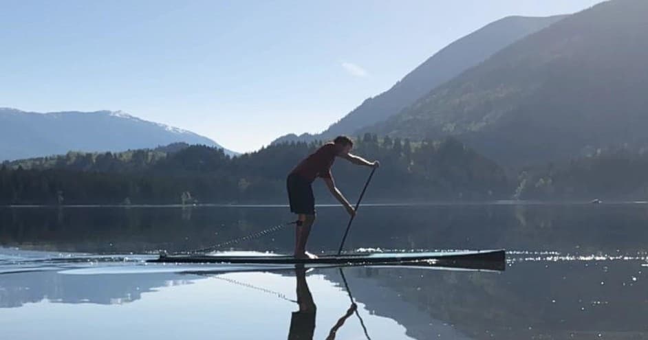 Stand-up paddleboarder gliding on reflective mountain lake at sunrise