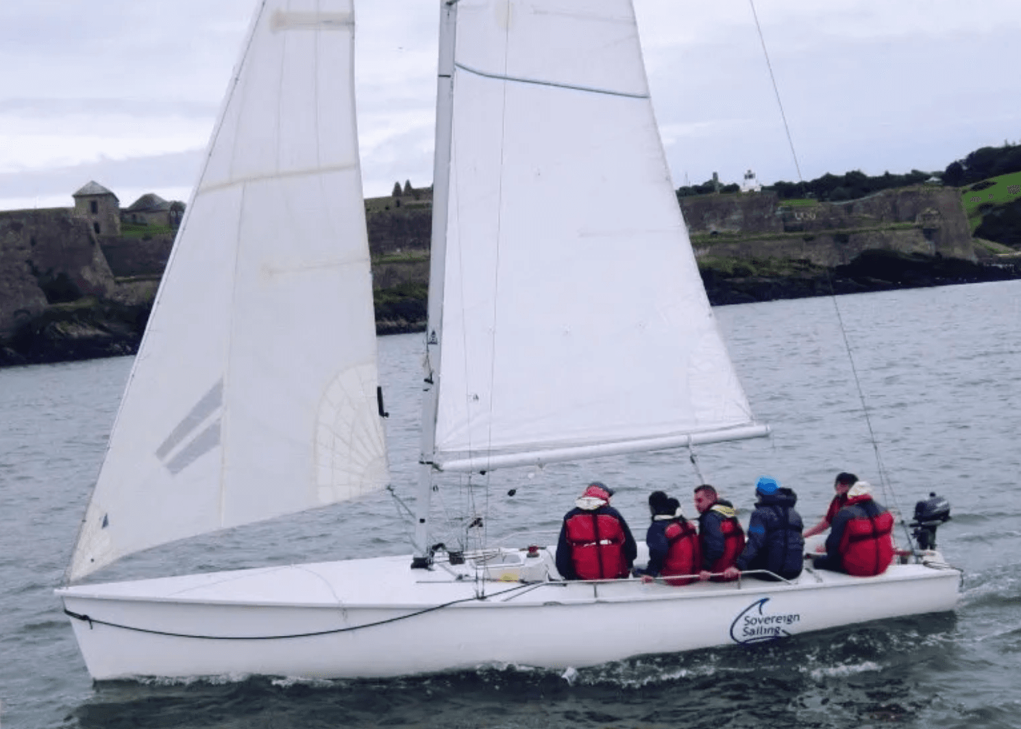 Group sailing lesson on small keelboat near rocky coast, training