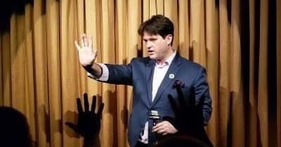 Man speaking on small indoor stage with audience hands raised